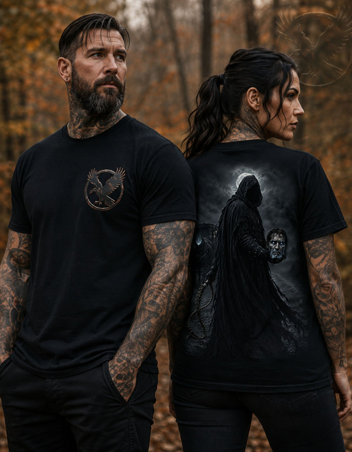 Two tattooed adults standing outdoors in a forest wearing black graphic T-shirts. The man on the left shows a small circular bird emblem on the chest, and the woman on the right shows a large back print of a hooded dark figure against a smoky sky.