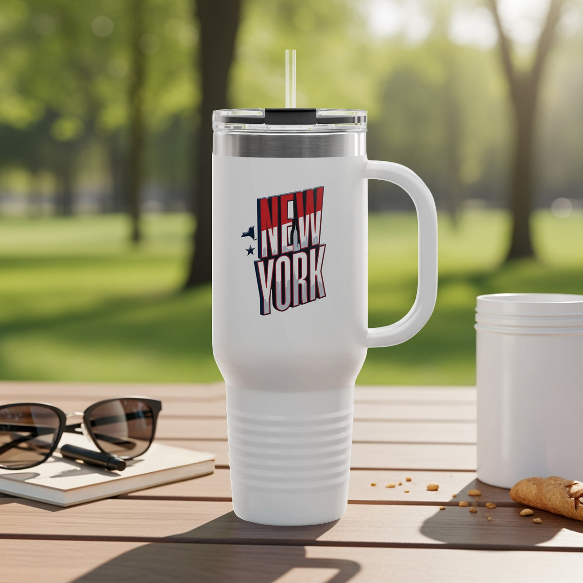 White travel mug with "New York" logo, sunglasses, and croissant on a table.