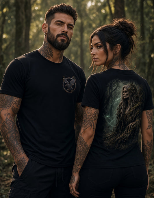 Two tattooed adults standing in a forest wearing black graphic T-shirts. The man on the left wears a small dark chest emblem, and the woman on the right shows a large back graphic of a ghostly figure in misty green and gray tones.