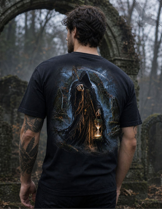 A man viewed from behind wearing a black graphic t-shirt with a detailed illustration of a hooded figure holding a glowing hourglass in a graveyard setting. The background is a blurred, misty forest cemetery.