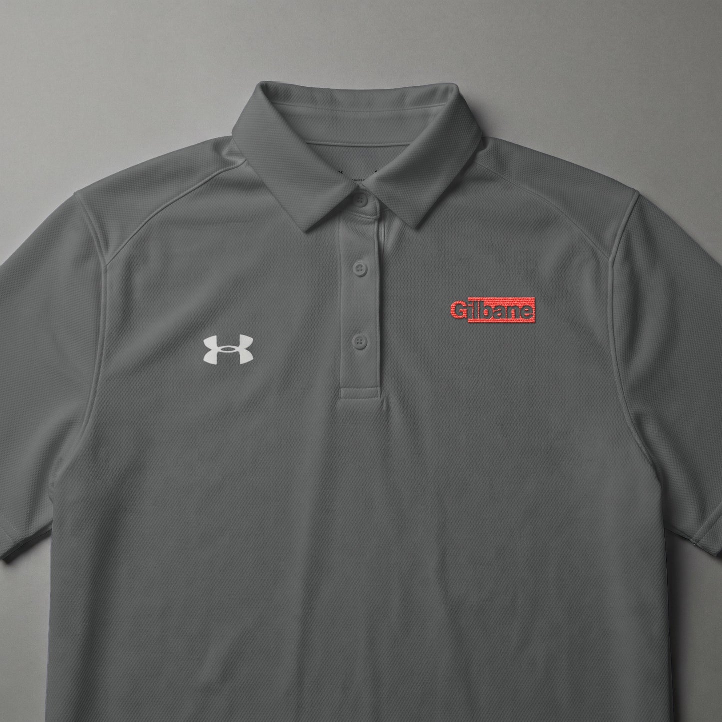 Grey Under Armour polo shirt with red Gilbane logo on chest.