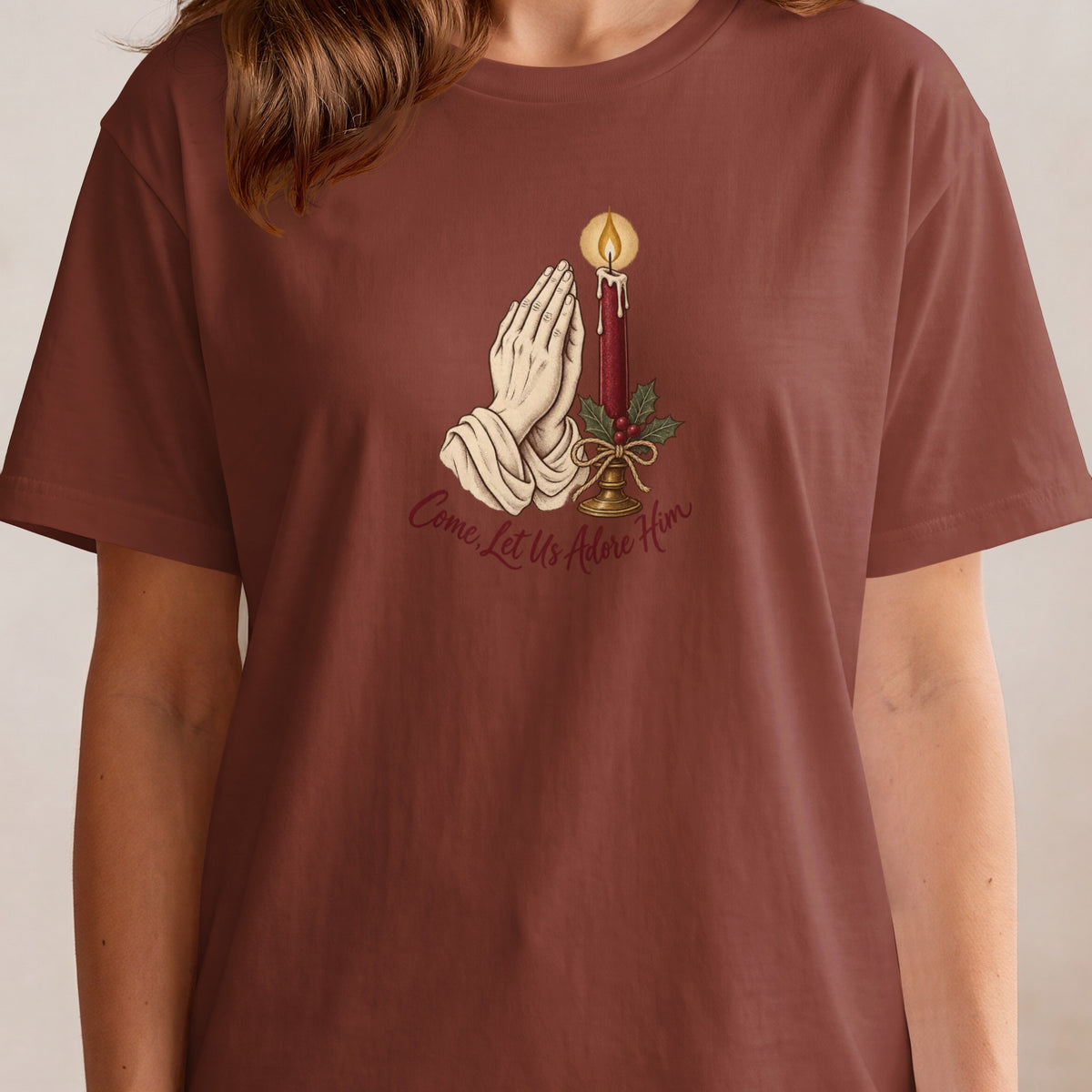 Praying hands, candle, holly, and text "Come, Let Us Adore Him" on brown shirt.