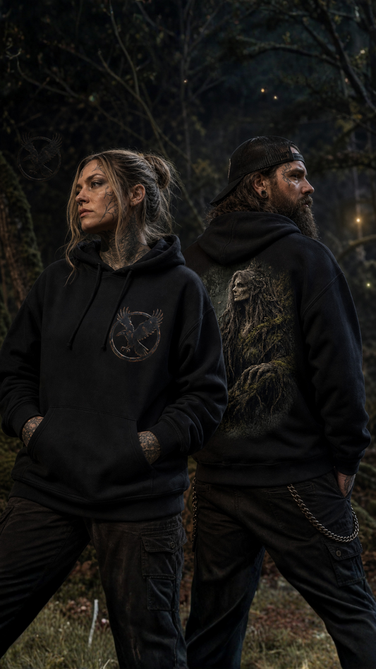 Two people standing outdoors in a dark forest setting, both wearing black hoodies. The person on the left wears a black hoodie with a circular bird logo on the chest, and the person on the right wears a black hoodie with a large detailed graphic on the back.