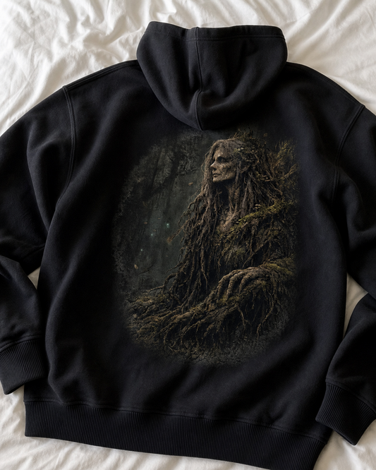 A black pullover hoodie laid flat on white bedding, showing a large back graphic of a mossy, tree-like female figure in a dark forest scene.