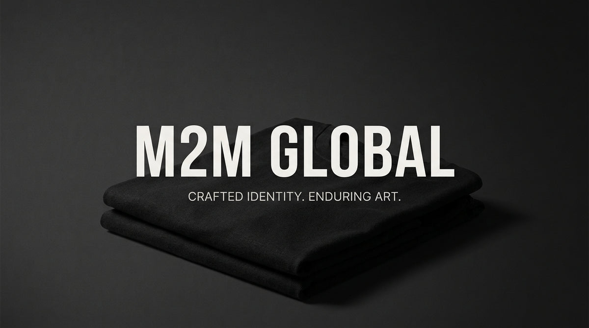 Folded black fabric, likely a bandana or scarf, displayed on a dark gray background with bold white text reading 'M2M GLOBAL' and smaller text 'CRAFTED IDENTITY. ENDURING ART.