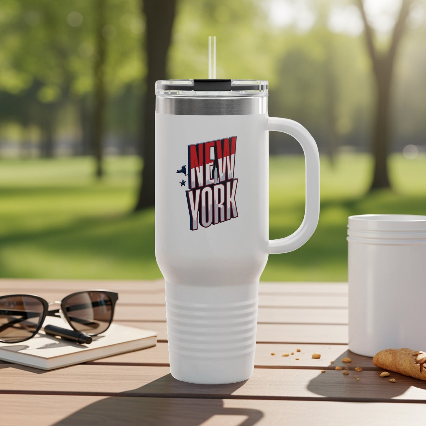 White travel mug with "New York" logo, sunglasses, and croissant on a table.