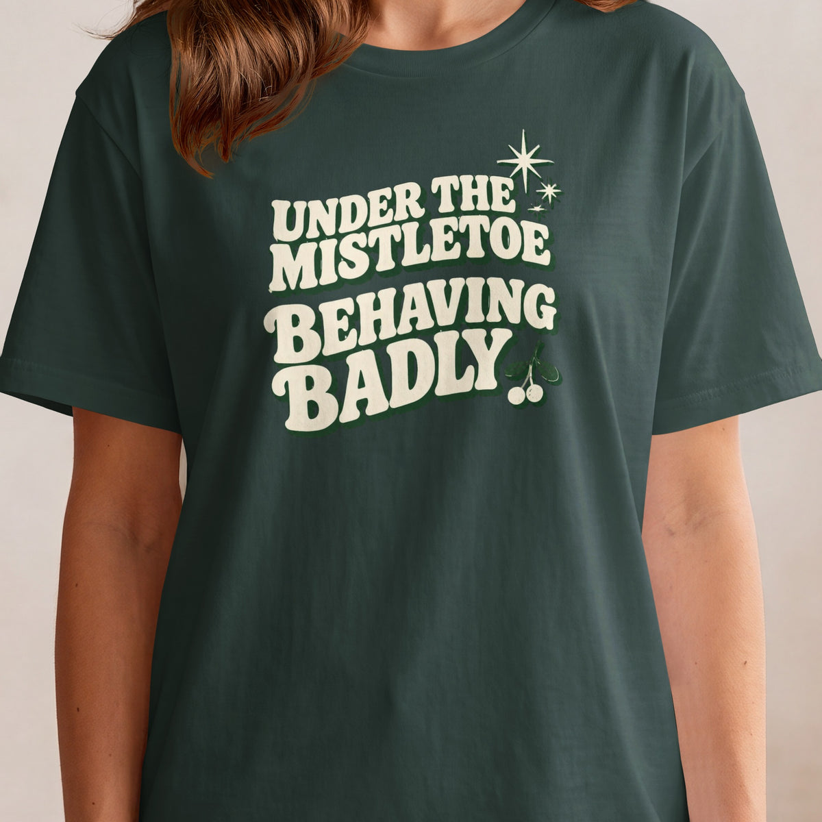 Dark green t-shirt with "Under the Mistletoe Behaving Badly" text and mistletoe sprigs.